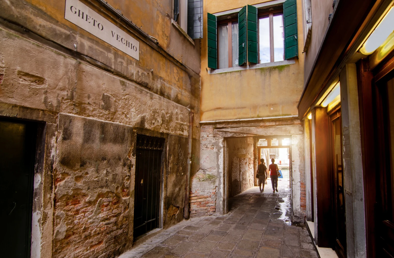 Two people walking toward daylight through a narrow passage between weathered buildings, photographed by Massimo Parisi.